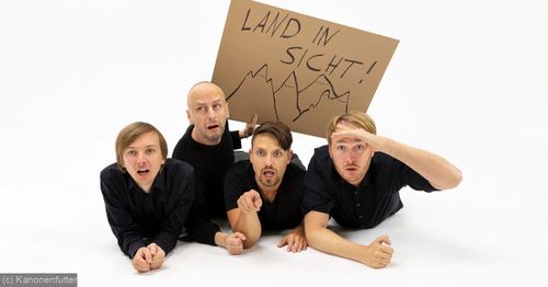 Vier Schauspieler liegen auf dem Boden und blicken überrascht nach vorne. Einer hält ein Papp-Plakat mit einem Gebirgsprofil in die Höhe auf dem zu Lesen steht: Land in Sicht!